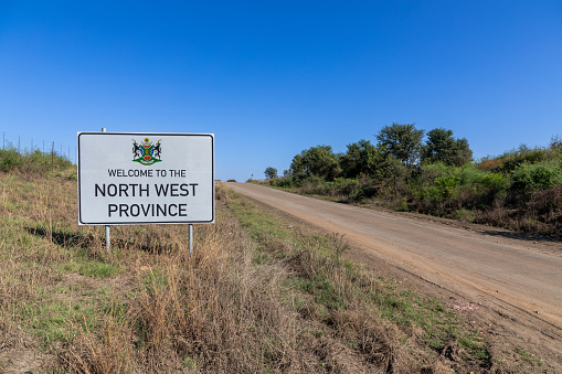 Welcome to North West Province Sign with a gravel road to the right of the sign .