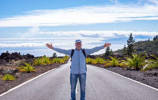 Active elderly man with backpack and open arms walking on the mountain road enjoys the beauty of nature in Tenerife, sea horizon, La Gomera island and wonderful blue sky