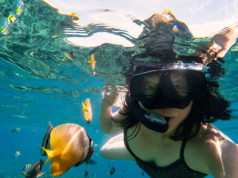 A solo senior Chinese woman takes a selfie while snorkeling underwater with tropical fish in Amed, Bali, Indonesia.