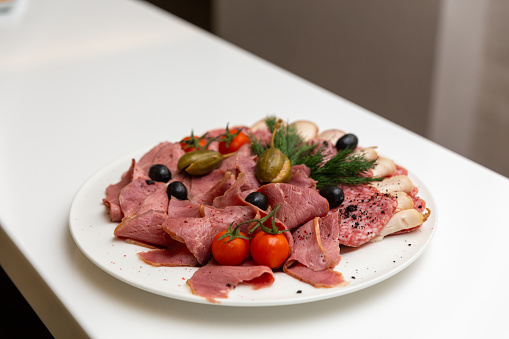 Meat delicatessen plate arranged with cherry tomato, black and green olives and capers. Set of sliced meat on a white plate.