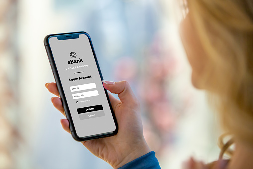 Observe the simplicity as a woman interacts with her mobile phone, accessing the secure login screen of the e-bank app. Experience the ease of managing finances and accessing account details with modern banking technology.