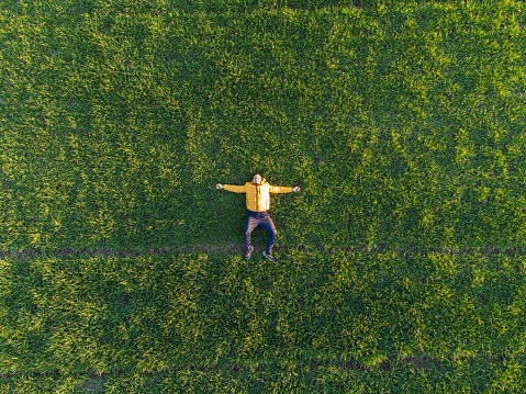 Young adult man in a yellow jacket is lying in a meadow of green grass. Feeling happy, carefree, enjoying outdoors time, enjoying spring, freedom concept.