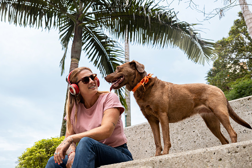 A woman, wearing red headphones and sunglasses, sits while looking at her pet with palm trees in the background.