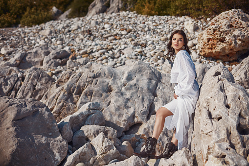A woman in a white dress leaning against a rock in a rocky area.