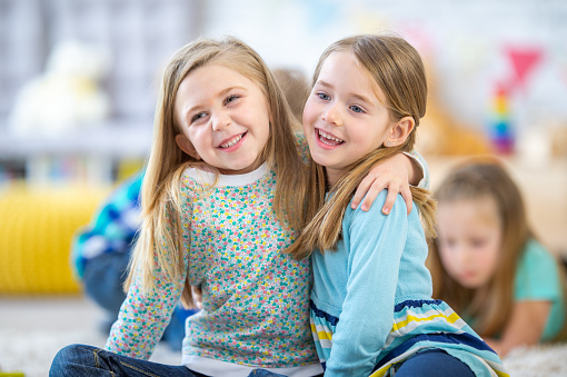 Two little girls sit on the floor of their kindergarten classroom , with their arms around one another, as they pose for a picture. They are both dressed casually and are smiling.