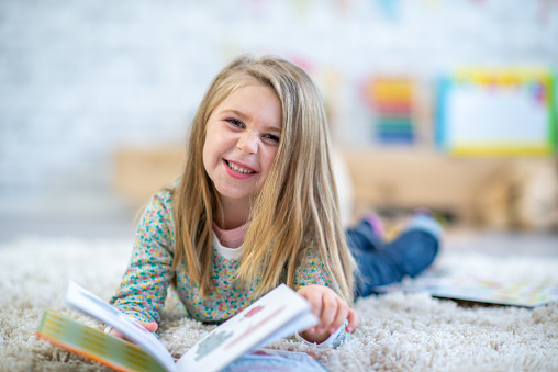 A sweet little blond-haired girl in kindergarten, lays on the floor of her classroom with a book open in front of her.  She is dressed casually and looking up from the story to smile.