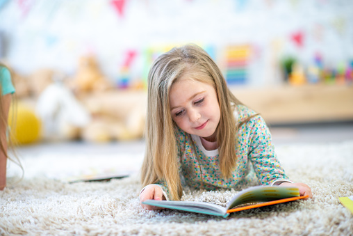 A sweet little blond-haired girl in kindergarten, lays on the floor of her classroom with a book open in front of her.  She is dressed casually and focused on reading the story.
