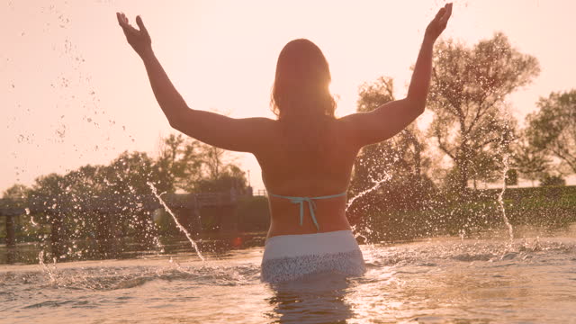 SLOW MOTION, LENS FLARE: Young female gracefully raises arms and splashes water