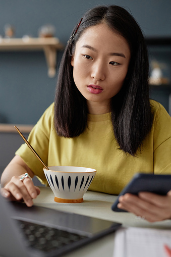 Vertical portrait of young Asian female freelancer holding smartphone working on laptop during breakfast