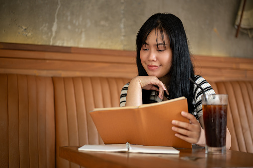 A happy young Asian woman in casual wear is reading a book or doing homework in a beautiful vintage coffee shop. people and lifestyle concepts
