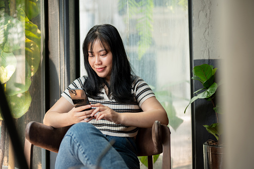 A positive Asian woman in casual clothes using her smartphone, chatting or scrolling on social media while sitting on an armchair in a beautiful green cafe. people and lifestyle concepts