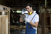 A carpenter or woodworker holding a tablet, reviewing design plan or instruction related to the woodwork. Wearing safety goggle, hearing protection headphone and glove stand in a woodworking workshop.