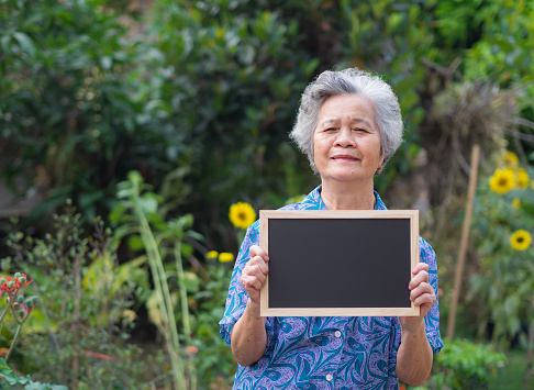 Happy Asian senior woman holding blackboard in the garden at home. Space for text.