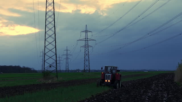 Drone Shot of Tractor Plowing Agricultural Field Under Power Lines and Cloudy Sky at Dusk
