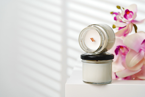 A small, opened jar of a scented candle sits on a white stand, its wick visible and unlit, with the soft glow of natural light filtering through blinds in the background. A delicate orchid, out of focus, adds a touch of elegance to the serene setting.