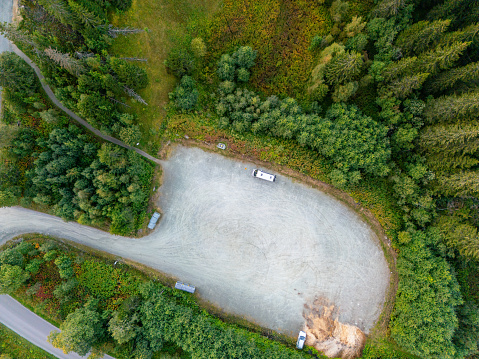 A top-view shows a gravel parking lot hidden within dense woods. A narrow road connects this area to main traffic. The presence of a few cars indicates it's a popular starting point for hikers or a rest stop for travelers. Variation in the trees' colors suggests different tree types or changing seasons. This secluded spot offers significant privacy and quiet.