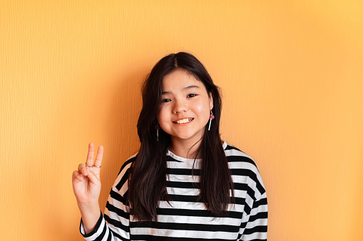 Sunny Disposition : A cheerful girl with a beaming smile makes a peace sign, set against a warm yellow backdrop that matches her sunny personality