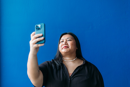 A fashionable woman takes a selfie in front of a blue concrete wall