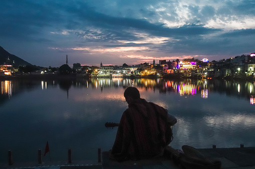 Young lonely guitarist playing on acoustic guitar in front of Pushkar Holy Lake during sunset twilight with lights reflecting on the lake over a beautiful dramatic sky