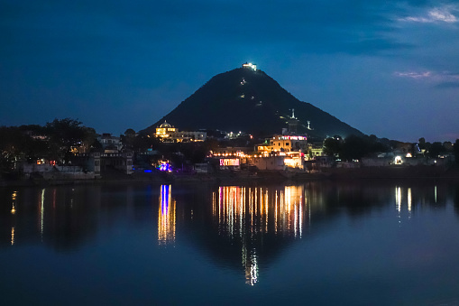 Savitri Devi Temple on top of a hill in front of Pushkar Holy Lake during twilight with beautiful city lights reflecting over the lake, is one of the sacred pilgrimage sites in Hinduism