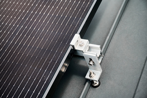 Close up of solar panel, securely fastened with robust mounting clamp to supporting structure on rooftop of house. Bolts and clamps provide stability and durability to solar installation.