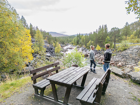 Two young people wearing traditional patterned sweaters, standing by a rocky river bank discussing close to an old picnic bench. The river moves slowly past, lined with trees, against a backdrop of rocks. This peaceful scene looks like a rest stop during a hike or road trip. In the far distance are mountains adding to the beauty of the scene.