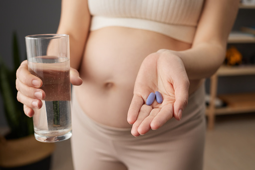 Unrecognizable pregnant woman with bare holding pill in her palm and glass of fresh water taking vitamins for future mothers while standing in living room at home