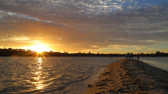 Tourists walking on the long sandbank (sandbar) stretching into the Swan River at Point Walter, Perth, Western Australia. Sunset, golden hour. Sun star, lens flare.