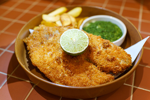 A plate of Fish and Chips is served with tartar sauce and mushy peas. The French fries are cut thickly in the British style, and a slice of lime rests on top of the fish.