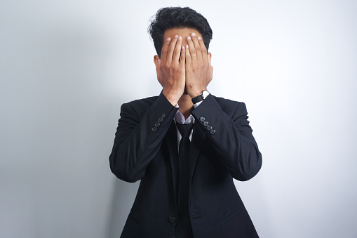 Man in suit covering his face with hands over white background.