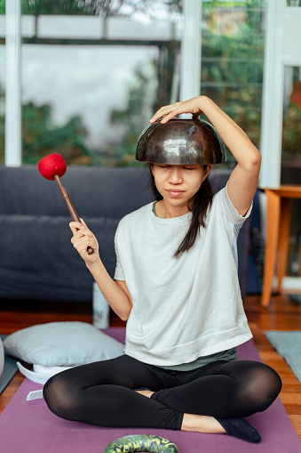 An Asian woman is using a Singing bowls for meditation that able to produce harmonic tones when struck or rubbed with a mallet, creating a calming and resonant sound with promoting mindfulness and reducing stress.