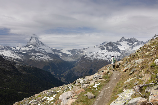 Female hiker walking on a small footpath in the Swiss alps, view of the Matterhorn in the background, overcast day.