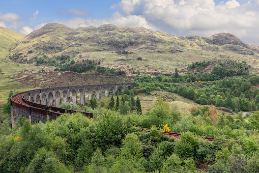The Glenfinnan Viaduct curves gracefully amidst verdant highlands, showcasing Scotland rugged terrain beneath soft, diffused sunlight
