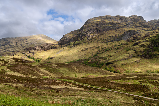 Verdant slopes of the Glen Coe valley, kissed by sunlight and shadows, with a winding trail offering a journey through the heart of the Scottish highlands