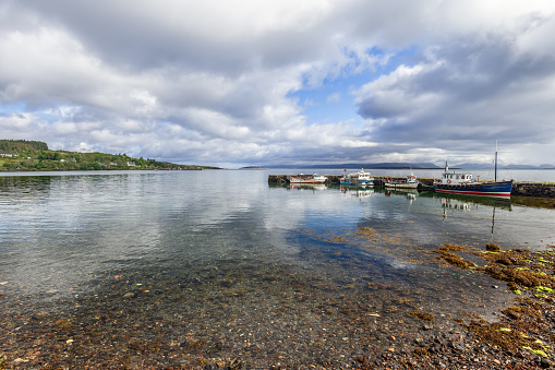 Morning light bathes the peaceful harbor of Broadford, showcasing moored vessels and a crystal-clear sea meeting the pebbled shore. Scotland