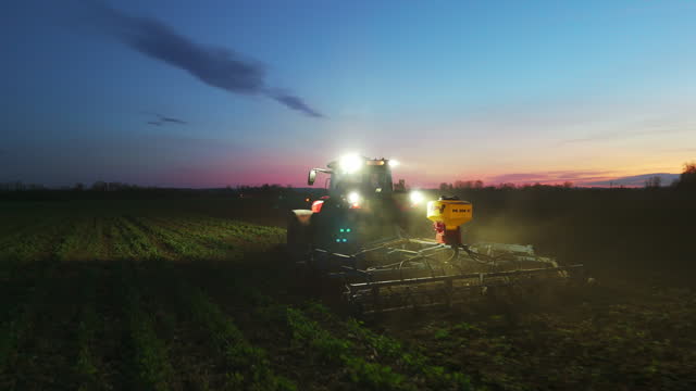 Illuminated Tractor Plowing Field Under Sky at Night in Village