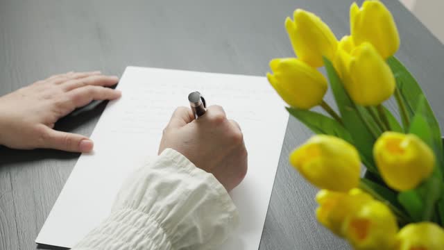 Hands of a woman writing a letter, story or report at a table decorated with yellow tulips.