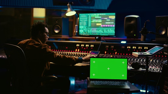 African american artist working with mockup on display in control room, mixing and mastering tracks with audio console and buttons or knobs. Music producer recording and editing sounds. Camera B.