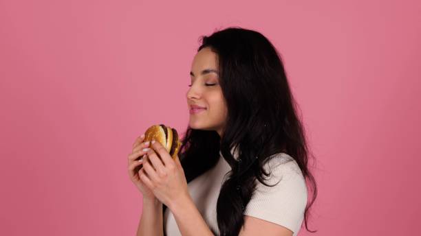istock Happy woman biting a hamburger while standing and posing in studio over pink background. People, lifestyle, junk food concept. Real time 2135054939