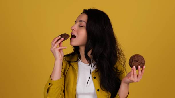 istock Happy young lady enjoying oat cookies while biting. Lady standing against yellow background. Healthy, junk food concept. Real time 2135055486