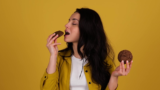 Smiling pretty woman holding two oatmeal cookies in hands and biting one, posing in studio, isolated on yellow background. Real time