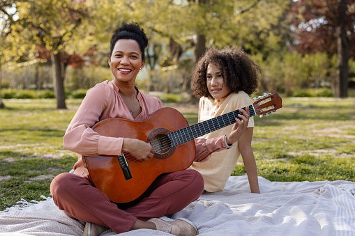 Happy african american mom and her teenage son playing the guitar while relaxing on a picnic in nature. Love, relationship and parenthood lifestyle concept