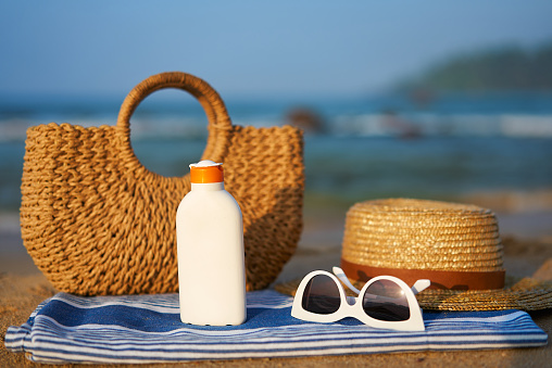 Summer accessories ready for day by sea, essentials for skin protection against UV rays. Straw bag, sun hat, sunglasses, sunscreen bottle on towel at sunny beach. Holiday scene with ocean backdrop.