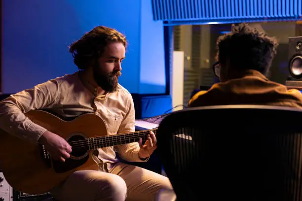 Singer playing electro acoustic guitar to record his tunes and compose a hit song, collaborating with audio technician. Young skilled artist performing music in professional studio, post production.