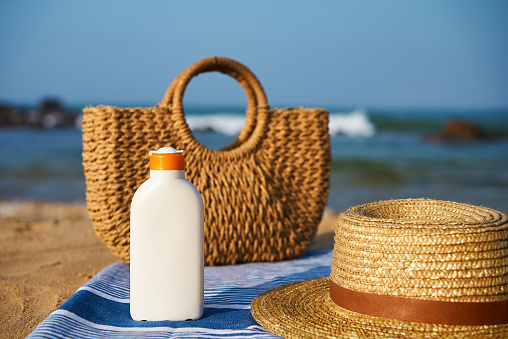 Summer accessories prepare for sunbath, promote skin safety, holiday essentials, beach day gear with ocean backdrop. Wicker bag, sun hat, sunscreen bottle on striped towel at sunny seaside.