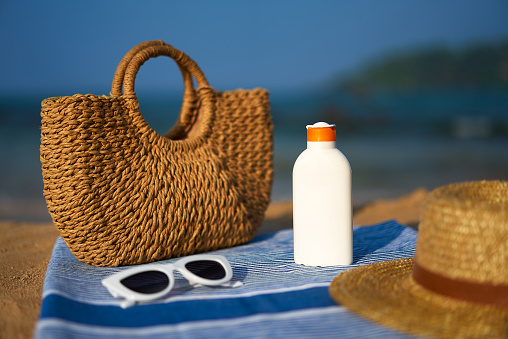 Items for safe sun exposure during seaside vacation. Straw bag, sunscreen bottle, beach, pair of white sunglasses rest on blue towel against sea backdrop. Essentials for day at beach under bright sun.