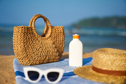 Bottle of sunscreen beside woven bag, sunglasses, sunhat on towel. Beach setup with sun protection essentials. Background of blue sea on sunny day. protection for skin health during seaside holiday.