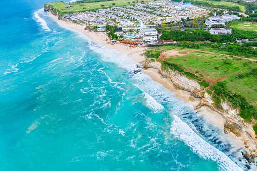 Bali, Bukit - The turquoise ocean waves crash against the eroded cliffside beach, showcasing the dynamic interaction between land and sea; adjacent lies a resort, characterized by organized housing structures.