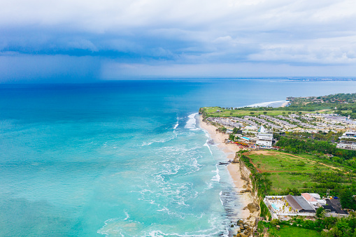 Bali, Bukit - The turquoise ocean waves crash against the eroded cliffside beach, showcasing the dynamic interaction between land and sea; adjacent lies a resort, characterized by organized housing structures and green fields, under a cloudy sky.
