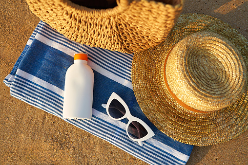 Sun protection for skin care on summer vacation. Sunscreen bottle on striped towel next to straw hat and sunglasses on sandy beach with ocean hint. UV rays block with SPF sunblock for safe tan.